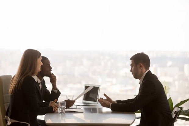 Friendly attentive hr managers interviewing vacancy applicant, multi-ethnic partners discussing new project idea sitting at office desk with cityscape outside, big window at background, side view