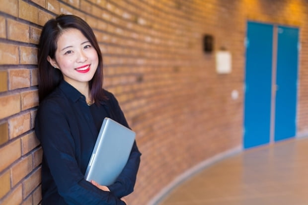 A portrait of an Asian college or university student at campus. She is standing at a red brick wall holding her laptop.