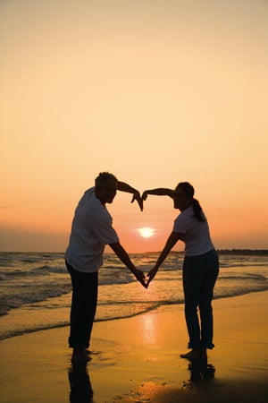 Mid-adult couple making heart shape with arms on beach at sunset.