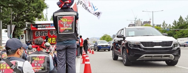 10일 오후 서구 기아 광주공장에서 생산된 번호판 없는 차량들이 결의대회 중인 화물연대 조합원들을 지나 다른 차고지로 이동하고 있다. 화물연대 파업으로 완성차를 운반하는 화물차량인 ‘카 캐리어’ 동원이 어렵자 기아는 임시방편으로 사무직원이 완성차를 직접 운전해 옮기도록 하고 있다.  /연합뉴스 